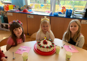 5 urodziny Ani. Ania z dwiema koleżankami siedzi przy stole, na którym stoi tort.